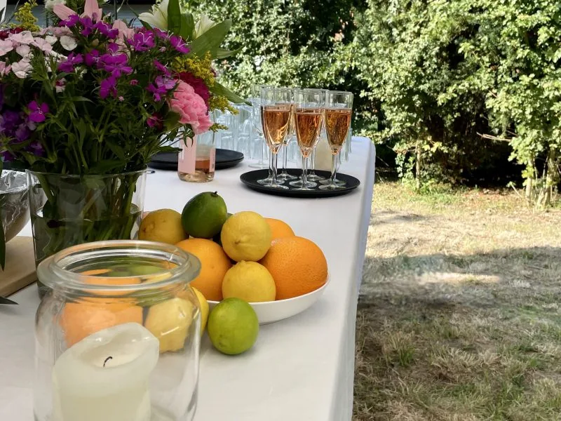 Tisch im Freien mit Blumenstrauß, Schale mit Zitrusfrüchten, Kerze im Glas und gefüllten Sektgläsern.