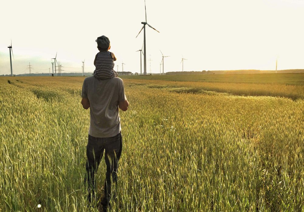 Mann mit kleinem Kind auf den Schultern steht in einem Getreidefeld und schaut in die Ferne auf Windräder