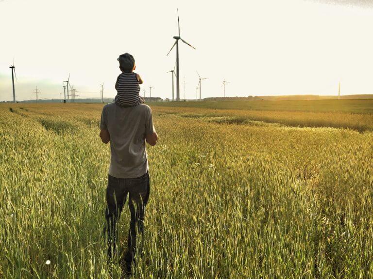 Mann mit kleinem Kind auf den Schultern steht in einem Getreidefeld und schaut in die Ferne auf Windräder