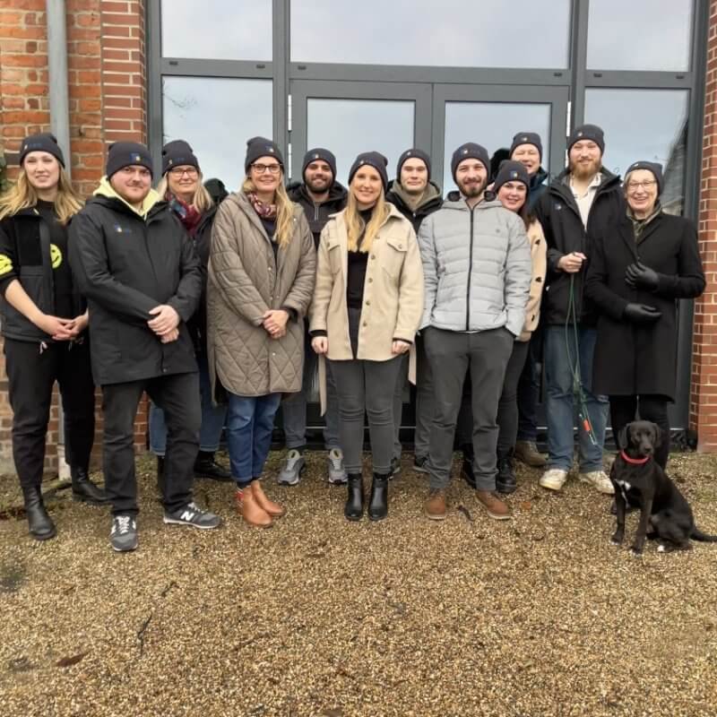 Mitarbeiter-Gruppenfoto vor dem Firmengebäude mit Strickmützen und Hund.